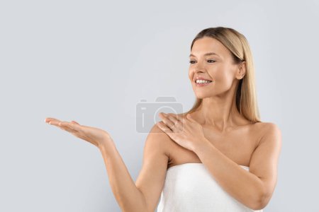 Foto de Cheerful attractive blonde middle aged woman wrapped in towel showing something invisible on her hand and touching shoulder, isolated on studio background. Body care cosmetics concept - Imagen libre de derechos