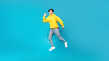 Photo for Joyful Chinese Teen Boy Jumping Smiling To Camera Posing Over Blue Background In Studio. Teenager Guy Celebrating Success Leaping In Mid Air Wearing Casual Clothes. Panorama, Full Length Shot - Royalty Free Image