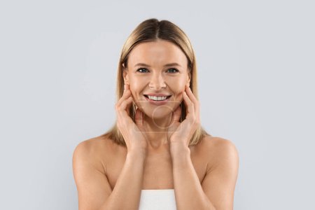 Foto de Face injections concept. Pretty blonde middle aged woman wrapped in white bath towel touching her face skin and smiling, enjoying results of face care treatment, grey studio background - Imagen libre de derechos