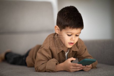 Foto de Busy serious european small kid plays on phone, watches video, lies on sofa in living room interior. App for meeting remotely, entertainment and communication, study, education with device at home - Imagen libre de derechos