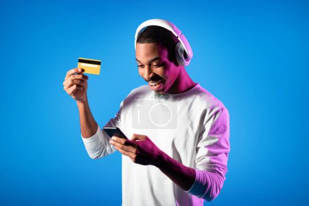 Téléchargez les photos : Newest musical app concept. Happy handsome millennial black guy in white longsleeve using modern cell phone, wireless headphones and yellow credit card over studio background in neon light - en image libre de droit