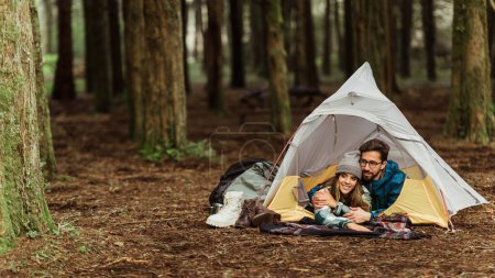 Téléchargez les photos : Cheerful millennial caucasian family in jackets resting in cold forest, enjoy adventure, lie in tent outdoor, panorama. Vacation, active lifestyle and camping, love, relationships and hikers sport - en image libre de droit