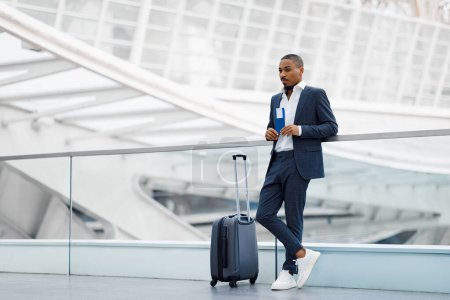 Foto de Portrait Of Young Black Businessman With Suitcase Waiting At Airport, Handsome African American Man Holding Passport With Tickets And Looking Away While Standing At Terminal, Copy Space - Imagen libre de derechos