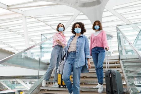 Foto de Tres amigas jóvenes que llevan máscaras médicas posando en las escaleras del aeropuerto, grupo de mujeres con maletas esperando el vuelo en la terminal, disfrutando de viajes seguros durante la pandemia del Coronavirus - Imagen libre de derechos