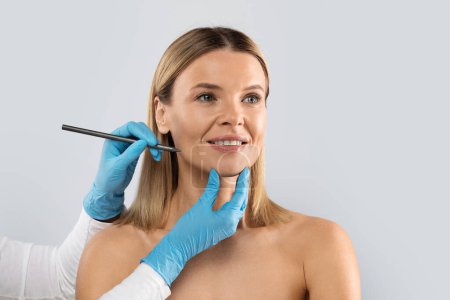 Photo for Hands of plastic surgeon wearing blue medical gloves drawing contours with black pen around female patient face before surgery, middle aged blonde woman getting beauty treatment, copy space - Royalty Free Image