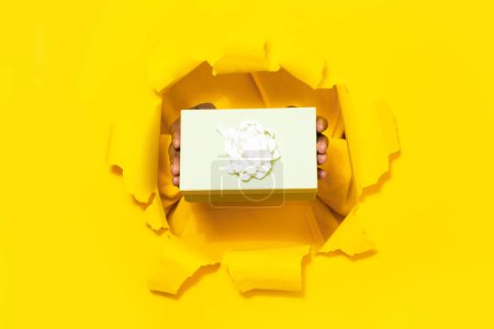 Foto de Male hands giving wrapped gift box through torn yellow paper background, closeup, free space. Present in package with ribbon bow. Special offer or sale concept - Imagen libre de derechos