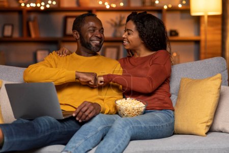 Joyful happy loving black couple sitting on couch in cozy living room, using modern laptop, eating popcorn, gambling or betting online, celebrating success, giving each other fist bump, copy space