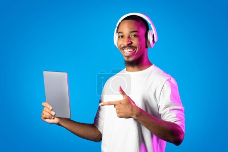 Photo for Cheerful handsome african american young man using digital pad and wireless headphones, listening to music on neon background, playing video games, watching video content, copy space - Royalty Free Image