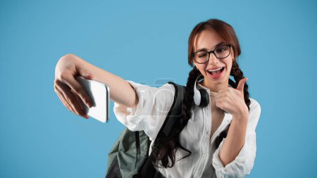 Téléchargez les photos : Adolescente caucasienne souriante avec des nattes dans les lunettes, l'élève prend selfie sur smartphone, montrer pouce vers le haut, isolé sur fond bleu, studio. Application photo pour l'éducation, blog et réseaux sociaux - en image libre de droit