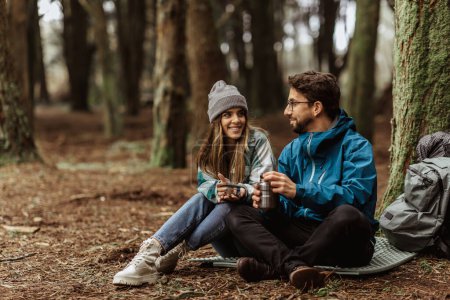 Photo for Glad young european guy and woman tourists in jackets with backpack rest in forest camping, enjoy cold season outdoor and cup of hot drink from thermos. Relax, date, lifestyle travel and relationships - Royalty Free Image