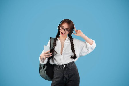 Photo for Happy caucasian teenager girl with pigtails in glasses with headphones student with backpack uses phone, sings and listens music, isolated on blue background, studio. Audio app for relax, education - Royalty Free Image