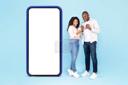 Téléchargez les photos : Excited black spouses standing near big smartphone with empty white screen, using gadget and shaking clenched fists, celebrating win over blue studio background, full body length collage - en image libre de droit