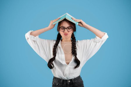 Upset caucasian teenager girl with pigtails in glasses female student presses book to head, suffering from overwork and study, isolated on blue background, studio. Problems with education, knowledge