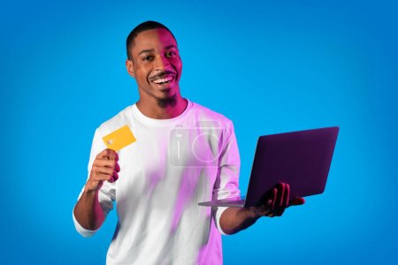 Happy bearded handsome young african american man in white holding modern laptop and credit card over neon studio background, smiling at camera, shopping on Internet, purchasing goods, copy space