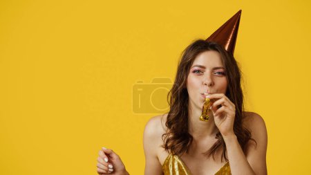 Téléchargez les photos : Jolie femme caucasienne en chapeau d'anniversaire soufflant corne de fête, célébrant sa journée spéciale sur fond de studio jaune, panorama avec espace libre. Jeune femme ayant plaisir vacances - en image libre de droit