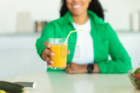 Photo for Healthy Drink. Unrecognizable Black Woman Offering A Glass Of Juice To Camera Sitting At Table In Kitchen At Home. Hydration And Vitamins Concept. Cropped, Selective Focus On Glass - Royalty Free Image