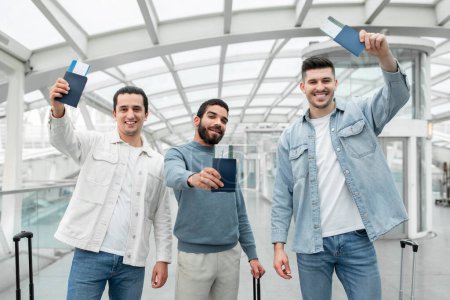 Téléchargez les photos : Three Male Friends Traveling Showing Boarding Pass Tickets Smiling To Camera Standing With Suitcases In Modern Airport Terminal Indoors. Happy Vacation With Friends Concept - en image libre de droit