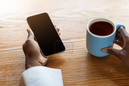 Foto de Female hands holding brand new cell phone with black empty screen and coffee mug, cropped of black woman sitting at table, chilling at cafe, copy space, mockup for online offer or mobile app ad - Imagen libre de derechos