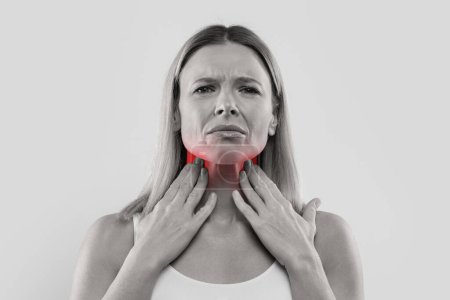 Foto de Sad blonde middle aged woman suffering from sore throat, rubbing red zones on her neck, black and white photo, isolated on studio background, closeup, copy space - Imagen libre de derechos