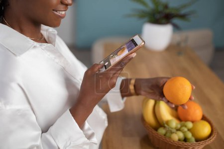 Foto de Cropped of black woman taking photo of bowl of fresh juicy orange, standing by wooden table, using smartphone. Food blogger making content for blog about healthy diet, detox, copy space - Imagen libre de derechos