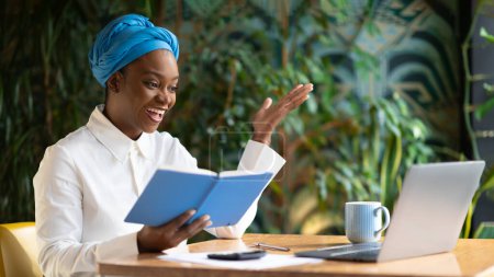 Photo for Cheerful beautiuful young african american woman in smart casual business advisor attending online business meeting, sit at table in front of laptop, holding notepad, cafe interior, copy space - Royalty Free Image