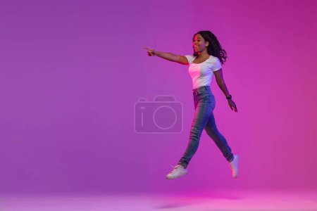 Téléchargez les photos : Look There. Happy Young Black Woman Jumping In Neon Light And Pointing Aside At Copy Space, Cheerful African American Lady Demonstrating Free Place For Advertisement On Purple Background, Full Length - en image libre de droit