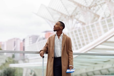 Téléchargez les photos : Young African American Man Waiting For His Flight In Modern Airport, Handsome Black Male In Stylish Coat Holding Passport With Ticket And Carrying Suitcase, Ready For Air Travel, Copy Space - en image libre de droit