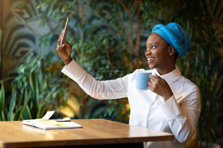 Téléchargez les photos : Attractive cheerful young african american woman in smart casual and blue headscarf chilling at cafe, sitting at table alone, drinking coffee, taking selfie on smartphone, copy space - en image libre de droit