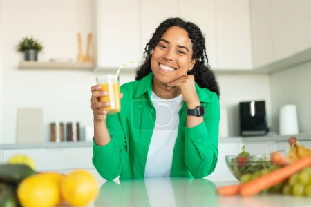 Téléchargez les photos : Une nutrition saine. Femme afro-américaine joyeuse buvant du jus d'orange frais tenant le verre et souriant à la caméra assise à la table à manger dans la cuisine moderne à la maison. Concept de régime de perte de poids - en image libre de droit