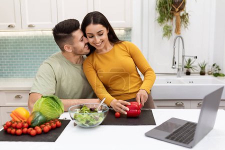 Photo for Cheerful attractive young couple standing by kitchen desk full of fresh organic vegetables, using modern computer, embracing, looking for nice recipe on Internet, copy space - Royalty Free Image