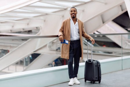 Foto de Hombre negro sonriente guapo que espera el vuelo en la terminal del aeropuerto, joven afroamericano de pie con la maleta, sosteniendo el pasaporte con boletos y mirando a la cámara, listo para el viaje, espacio de copia - Imagen libre de derechos