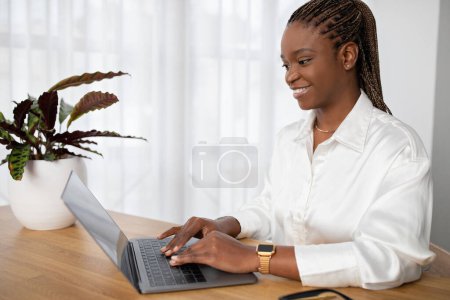 Foto de Feliz joven afroamericana de pelo bastante largo en traje formal sentada en el escritorio de la oficina, trabajando en el ordenador portátil, escribiendo en el teclado de la computadora y sonriendo, enviando correos electrónicos, espacio para copiar - Imagen libre de derechos