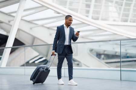 Photo for Portrait Of Handsome Black Businessman Walking With Suitcase In Airport And Using Smartphone, Young African American Man In Suit Browsing Internet On Cellphone While Going To Flight Gate, Copy Space - Royalty Free Image