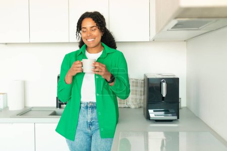 Téléchargez les photos : Cheerful African American Woman Drinking Coffee Enjoying Taste With Eyes Closed, Posing Holding Cup Standing Near Coffee Machine In Modern Kitchen Indoors On Weekend Morning - en image libre de droit
