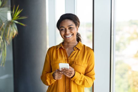 Téléchargez les photos : Portrait d'une heureuse femme d'affaires afro-américaine utilisant un smartphone, envoyant des SMS avec un client tout en restant près de la fenêtre dans son bureau, copiant de l'espace. Concept d'entreprise et de télétravail - en image libre de droit