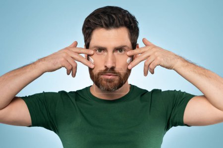 Photo for Handsome bearded man applying cream on his face, middle aged male with cream on his cheeks from wrinkles and dryness, blue studio background. Eye and face care for men - Royalty Free Image