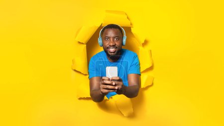 Téléchargez les photos : Homme afro-américain positif dans un casque sans fil, écoutant de la musique et utilisant un téléphone portable, debout à travers un trou dans du papier jaune déchiré, panorama. Offre musicale - en image libre de droit