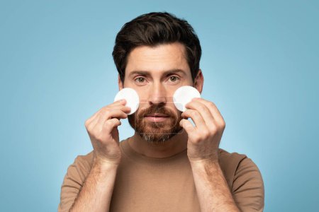 Foto de Handsome middle aged man cleaning face skin with cotton pads and looking at camera over blue background. Skin care and male beauty treatment - Imagen libre de derechos
