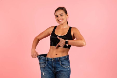 Photo for Happy Slim Woman Wearing Oversized Jeans And Pointing At Her Sporty Body, Smiling Young Sporty Female Demonstrating Weight Loss Result While Posing Isolated Over Pink Background, Copy Space - Royalty Free Image