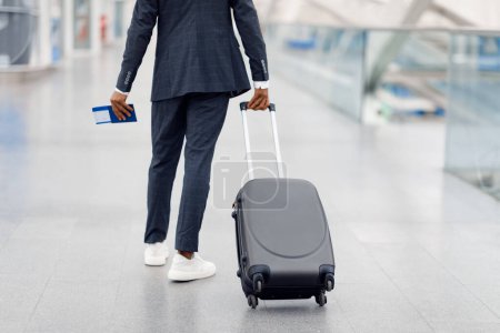 Téléchargez les photos : Homme noir méconnaissable portant un costume marchant avec une valise à l'aéroport, prise de vue recadrée d'un jeune voyageur afro-américain tenant un passeport avec des billets et allant à la porte d'envol, vue arrière - en image libre de droit
