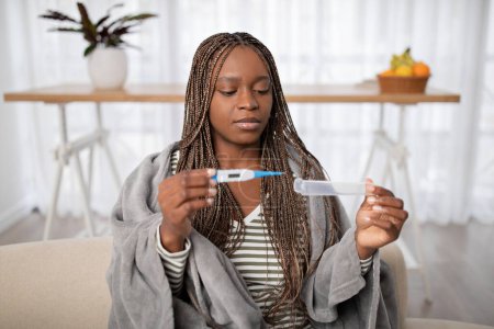 Photo for Unhappy pretty young black woman with long braids sitting on couch under warm eiderdown at home, looking at electronic thermometer in her hands, fever for no reason concept, copy space - Royalty Free Image