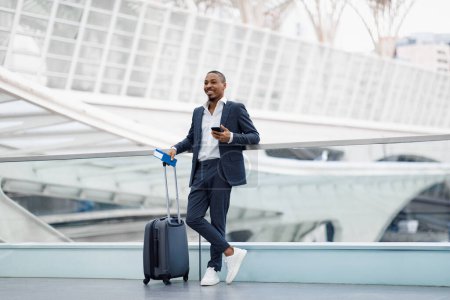 Téléchargez les photos : Bel homme d'affaires afro-américain debout avec valise à l'aéroport, portrait d'un jeune entrepreneur noir souriant tenant un passeport avec des billets et un téléphone intelligent, en attente d'un vol au terminal - en image libre de droit