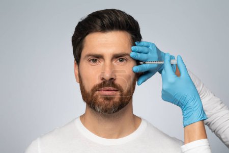 Photo for Handsome bearded man getting under eye injections in beauty clinic, looking at camera, standing on grey studio background. Male attending beautician, having face filler - Royalty Free Image