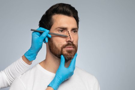 Téléchargez les photos : Docteur en gants de protection dessinant des marques sur le nez des hommes pour une opération de chirurgie esthétique, debout sur fond de studio gris, espace libre - en image libre de droit