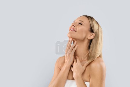 Foto de Smiling beautiful half-naked middle aged blonde woman covered in white towel touching her neck, enjoying anti-aging SPA salon treatment results, grey studio background, copy space - Imagen libre de derechos