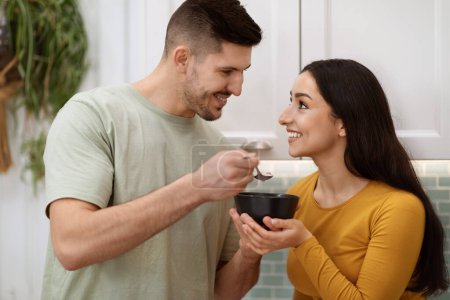Téléchargez les photos : Closeup of cheerful handsome young man feeding pretty happy girlfriend with long hair, husband giving wife spoon of cereals, granola or oatmeal, breakfast together, kitchen interior - en image libre de droit