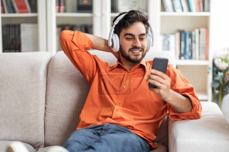 Photo for Positive happy handsome arab guy in casual outfit chilling on couch at home, middle eastern man sitting on sofa, using wireless headphones and cell phone, watching video content online, copy space - Royalty Free Image