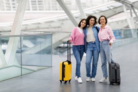 Téléchargez les photos : Portrait Of Three Happy Female Friends Posing At Airport, Cheerful Young Women Carrying Suitcases And Smiling At Camera While Standing At Terminal Hall, Group Of Ladies Enjoying Travelling Together - en image libre de droit