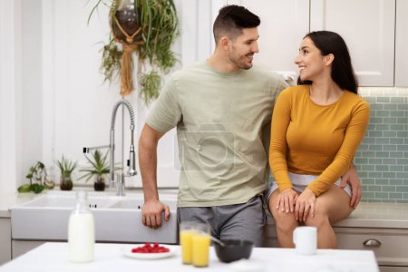 Téléchargez les photos : Loving beautiful young couple in homewear bonding while have breakfast together at home, happy man and woman drinking orange juice, eating granola, have conversation, smiling, copy space - en image libre de droit