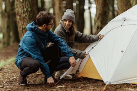 Téléchargez les photos : Smiling millennial caucasian guy and woman in jackets resting in cold forest, enjoy adventure, making tent outdoor. Vacation, active lifestyle and camping, hikers sports and walking at spare time - en image libre de droit